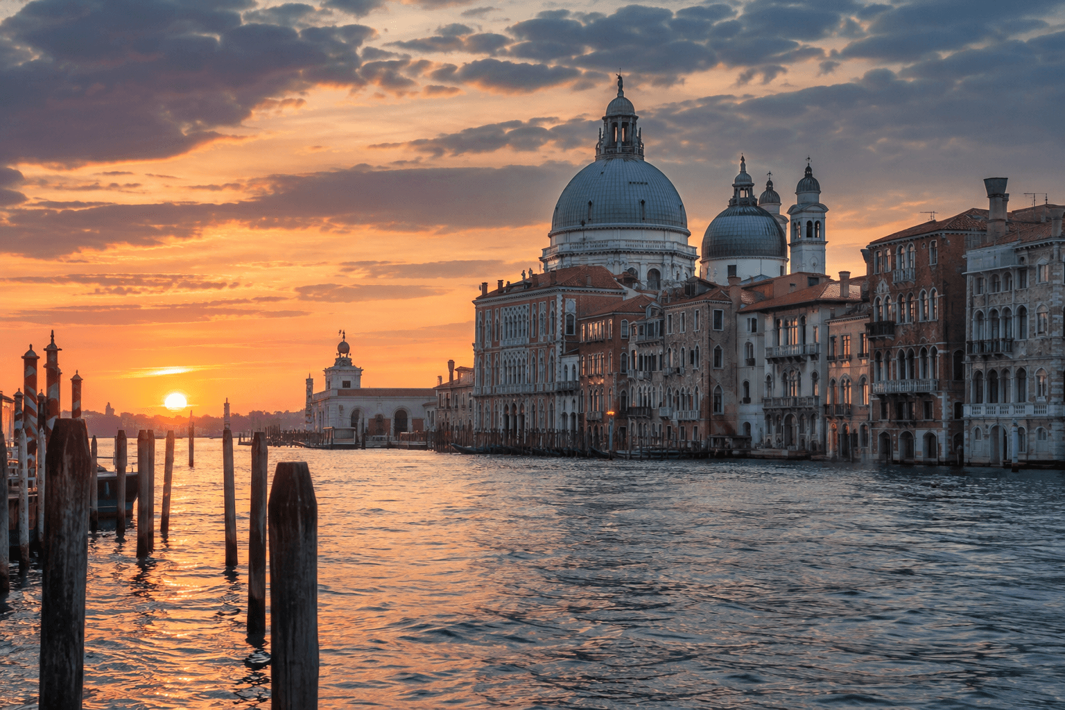 Illustrative view of the Grand Canal in Venice at sunset, with a domed basilica and wooden mooring poles in the water